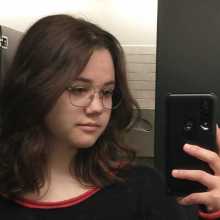 A mirror selfie of a person with brown shoulder-length hair and glasses, wearing a red and black sweater.