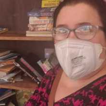 Brunette woman with glasses and wearing a mask poses in front of a messy pile of books.
