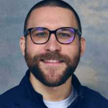 A picture of a 31 year old man, smiling, with glasses, short hair, and a beard.
