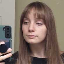 Girl with medium, straight brown hair and bangs. Taking mirror selfie with neutral expression.