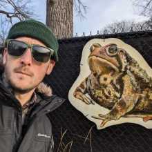A picture of Nick wearing a beanie and sunglasses and a parka, next to a large cardboard image of a frog