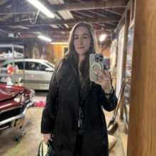 White woman in black trench coat takes selfie in garage