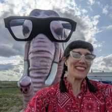 Photo of woman with brown hair and glasses wearing a red shirt and Sat ding in front of a statue of a pink elephant wearing glasses. 