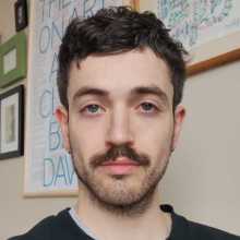 white man, dark hair, dark moustache, before a bedroom wall of framed pictures