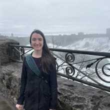 Caucasian women with long brown hair wearing black standing outside in front of a waterfall 