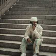 Me sitting on the stairs with a white cap and cream jacket and green cargo pants 