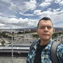 Un hombre con una camisa a cuadros verde y azul se toma una selfie. Detrás de él, hay una ciudad con edificios y montañas bajo un cielo nublado.