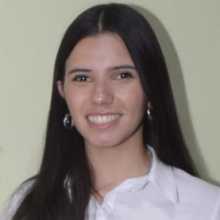 Mujer joven con el cabello largo y oscuro, sonriendo a la cámara, vestida con camisa blanca y posando frente a un fondo claro. Young woman with long dark hair, smiling at the camera, wearing a white shirt and posing against a light-colored background.
