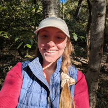 Woman with long braided hair, hat, and trees in the background