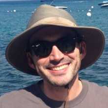 smiling caucasian man wearing a hat and sunglasses with a body of water in the background