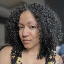 Person with curly dark hair, nose ring, glossy lips, wearing a black and gold patterned top and gold necklace, looking at camera with a slight smile.