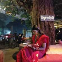 A girl reading a book in a sare under a big tree which has a sign saying free library in bangla