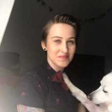 A white person with brown hair smiling in front of a darkened background, with a white fluffy dog in front of them laying one paw on their shoulder