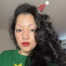 A smiling woman with long, dark curly hair wearing a green Christmas sweater and a red glittery Santa hat, festive holiday selfie.