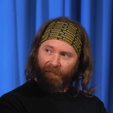 Man with headband sitting in front of blue curtain.