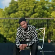 Person wearing a black jacket, sitting on a wooden chair with a green background.