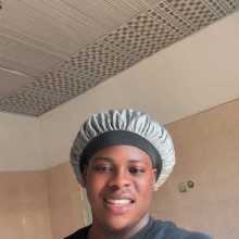 "A smiling young man wearing a black shirt and a grey silk hair bonnet, centered against a tiled wall and patterned ceiling.