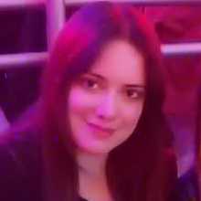 Person with long hair sitting indoors under pink and purple lighting.