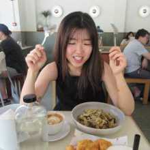 Girl with long hair holding a fork and spoon staring happily at the pasta dish in front of her in a cafe filled with people.
