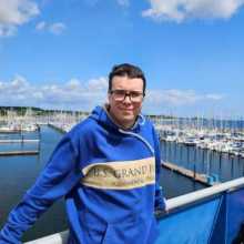 Man in late 20s in a blue sweatshirt with the blue sky sea panorama in the background