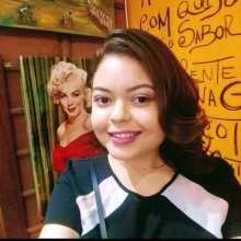 A young woman with wavy dark hair and a bright smile stands indoors, looking directly at the camera. In the background, there's a prominent painting of Marilyn Monroe in a red dress.