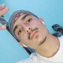 Young man wearing a patterned bandana and white t-shirt, making a peace sign, with a light blue wall in the background