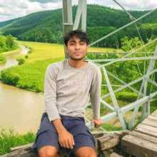 A young man wearing a light gray long-sleeve shirt and dark shorts is sitting on a wooden bridge with a green valley, winding river, forested hills, and cloudy sky in the background