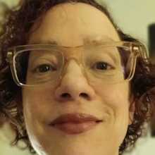 Headshot of a Caucasian woman with curly short hair, dark red lipstick, and pink colored clear glasses.