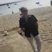 Well dressed gentleman, of average height, lightly muscular build, looking handsome on a beach, wearing a black chacabana shirt and white linen trousers. 
