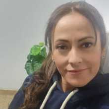 Woman with long dark wavy hair, smiling gently at the camera, wearing a navy blue hoodie, indoor setting with a green plant in the background.