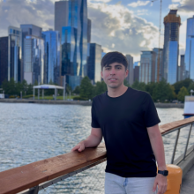 A young man wearing a black T-shirt and a cap, standing by the waterfront with a city skyline in the background